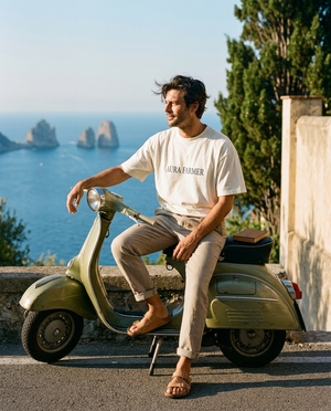 A man on a vintage olive-green Vespa pulled over on the Amalfi coast at golden hour, wearing the Aura Farmer cream cotton tee.