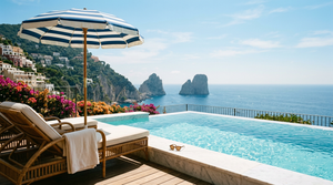 Private Capri pool deck with a vintage blue-and-white striped umbrella, rattan lounger, towel, and the Faraglioni rocks across the Tyrrhenian Sea.
